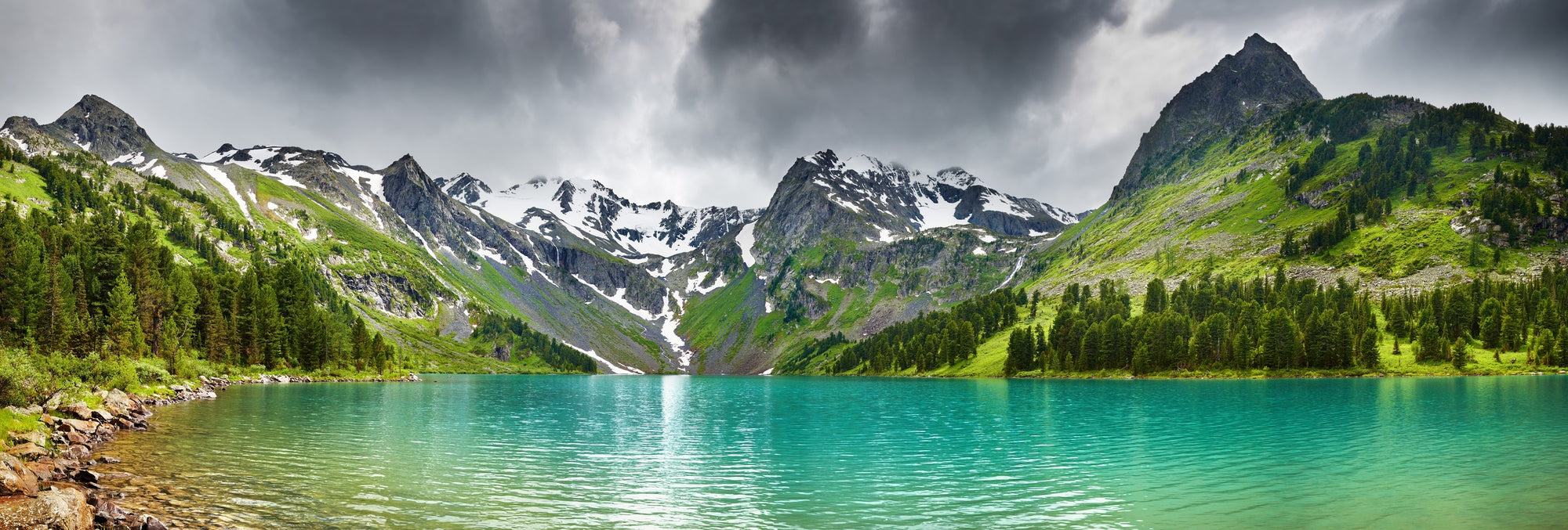 A aqua blue alpine lake is surrounded by green trees and snowcapped mountains.