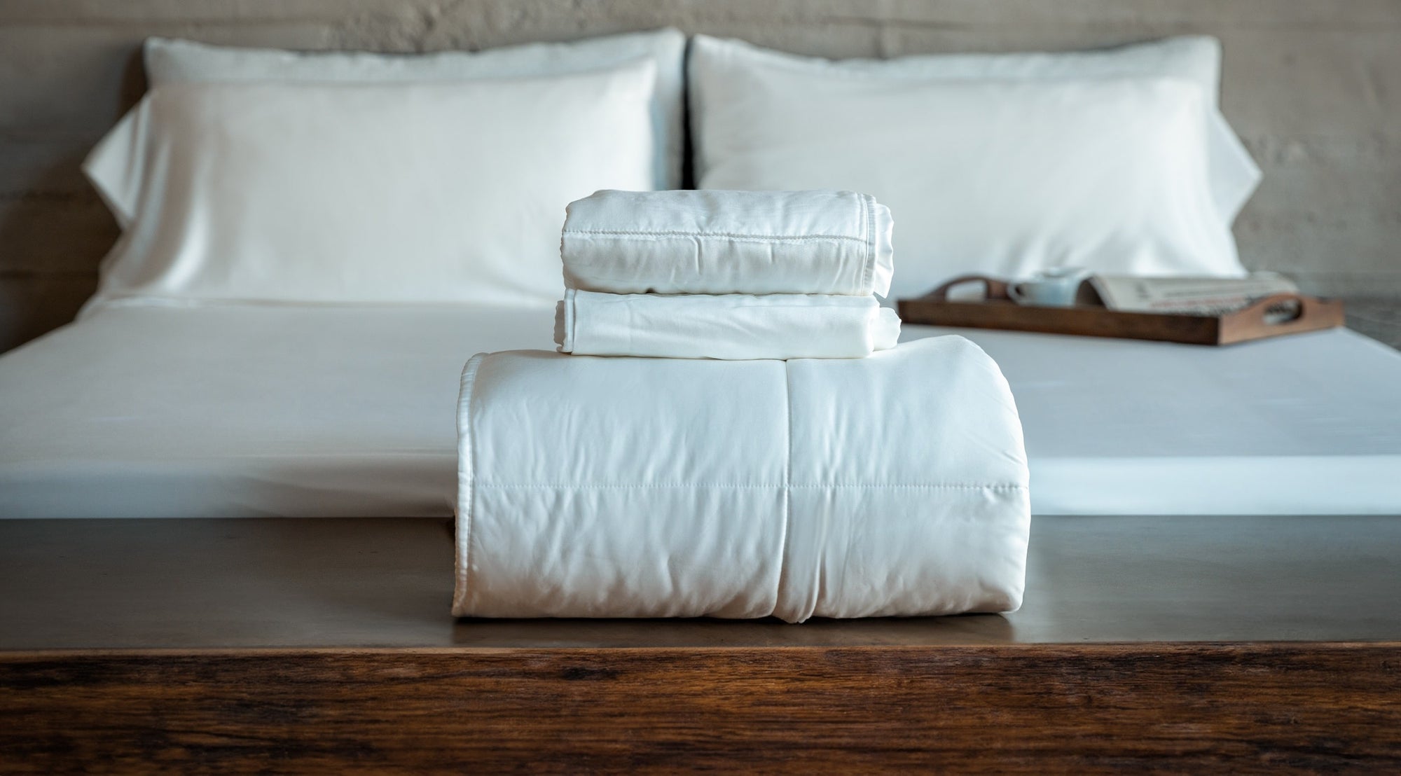 Sleepmod sustainable bedding products are stacked at the foot of a bed on a wooden table in a contemporary eco-friendly house.