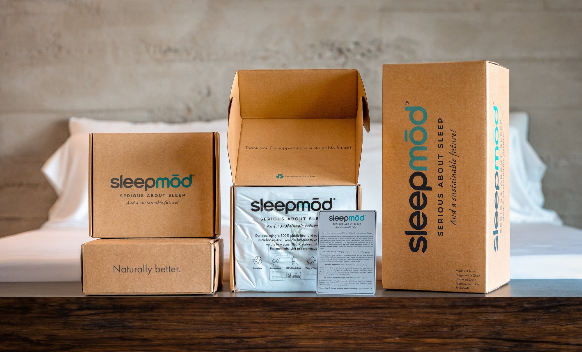 Sleepmod sustainable packaging on a warm wooden table in front of a bed made with a eucalyptus sheet set.