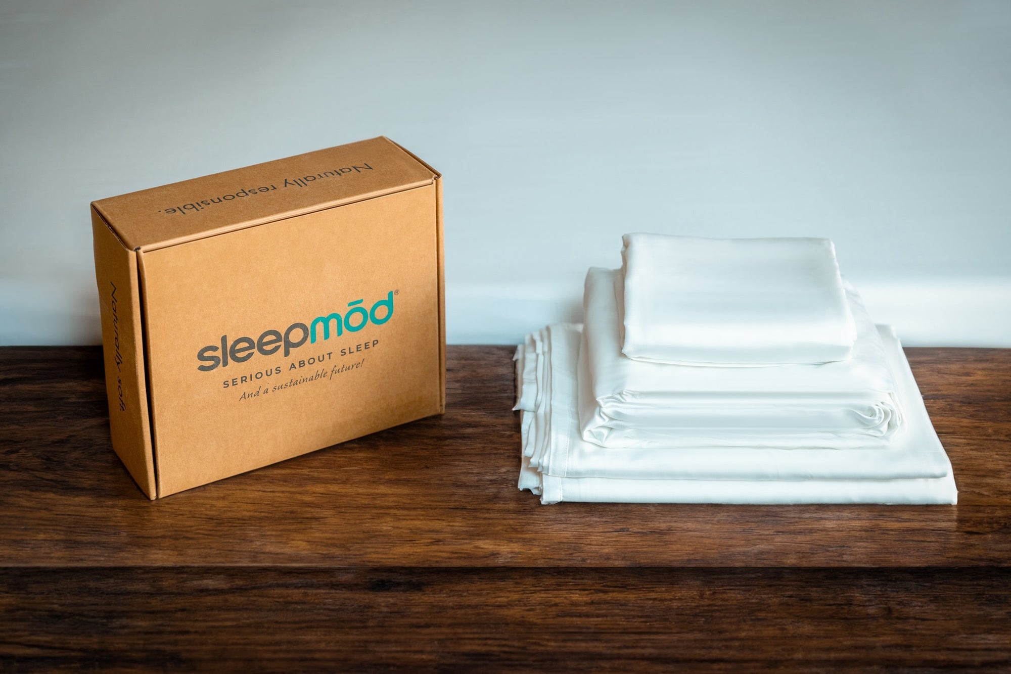 Sleepmod sustainable packaging on a warm wooden table in front of a bed made with a eucalyptus sheet set.