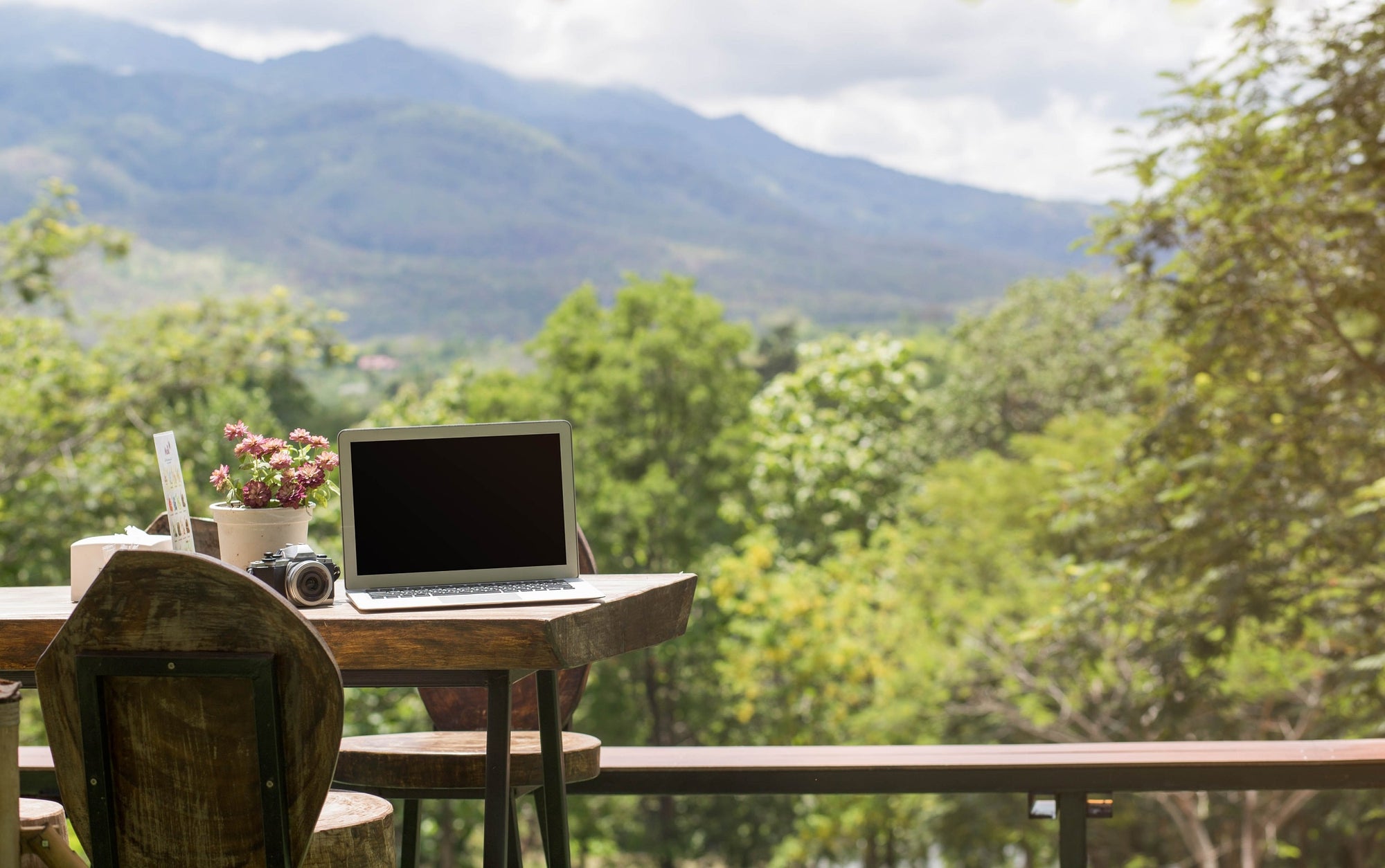 A laptop and camera sit on a sustainable raw wood table overlooking beautiful mountain greenery.